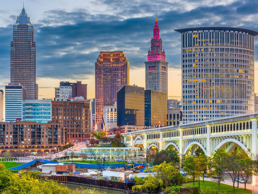 A vibrant cityscape at dusk in Ohio, featuring tall, illuminated buildings, a bridge in the foreground, and a partly cloudy sky.