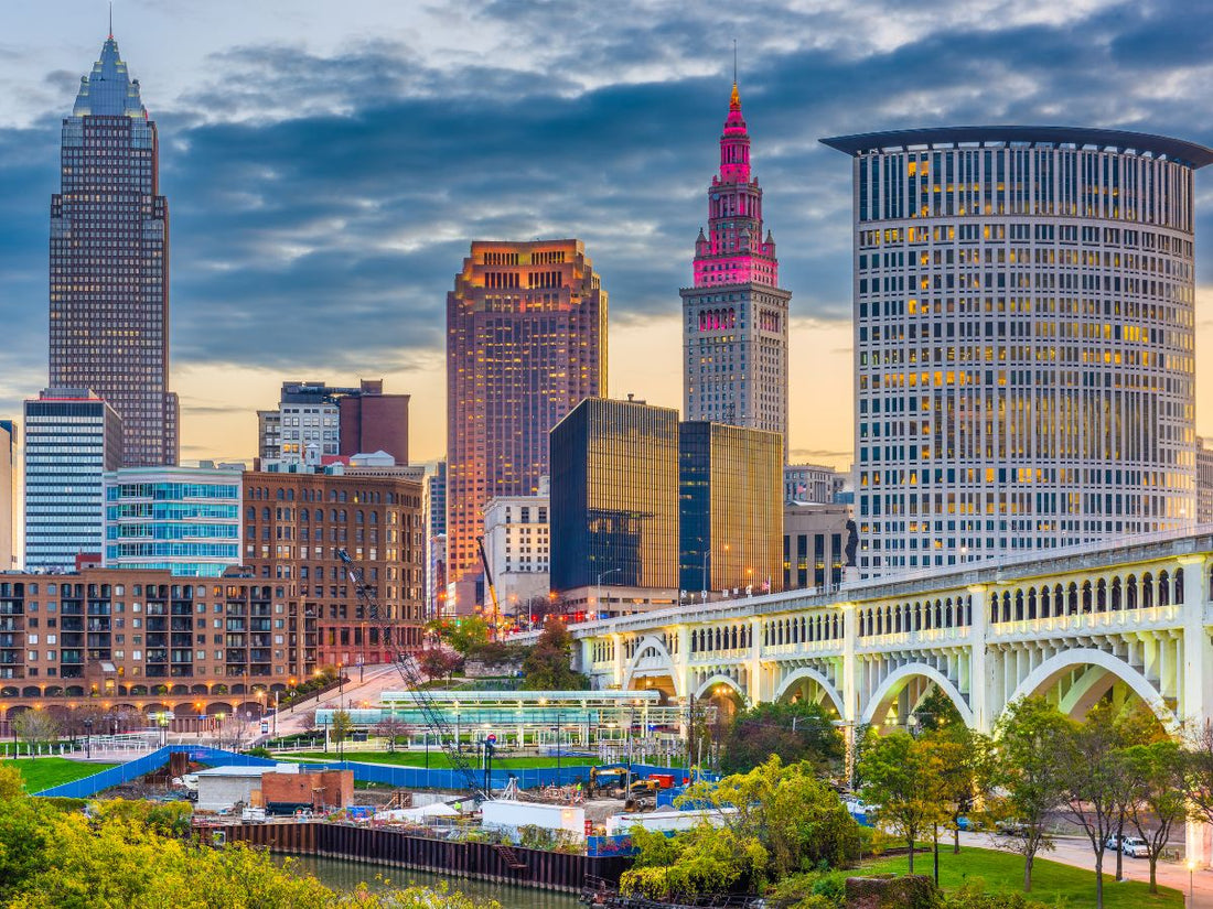 A vibrant cityscape at dusk in Ohio, featuring tall, illuminated buildings, a bridge in the foreground, and a partly cloudy sky.